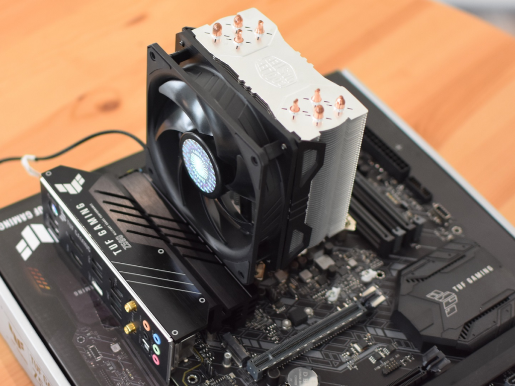 A professional, close-up shot of an air CPU cooler with its fan attached, mounted on a modern dark-colored motherboard, highlighting the heat pipes and aluminum fins against a blurred wooden background.