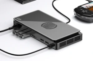 A sleek, modern external GPU enclosure in dark gray with a textured top panel, connected via a black cable to a portable gaming device, showcasing its compact size and connectivity ports. The enclosure features ventilation grilles on the side, suggesting efficient cooling for the internal components.