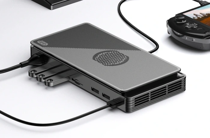 A sleek, modern external GPU enclosure in dark gray with a textured top panel, connected via a black cable to a portable gaming device, showcasing its compact size and connectivity ports. The enclosure features ventilation grilles on the side, suggesting efficient cooling for the internal components.